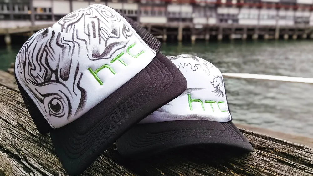 Two trucker hats featuring black hand-painted totemic linework and green HTC embroidery resting on weathered wood