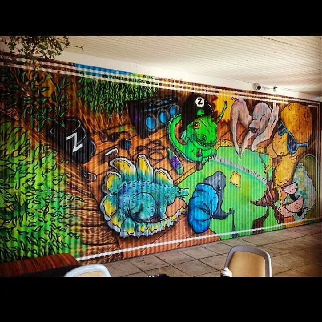 Mural on corrugated metal depicting vibrant cartoon dinosaurs, including a T-rex and stegosaurus, with a boombox