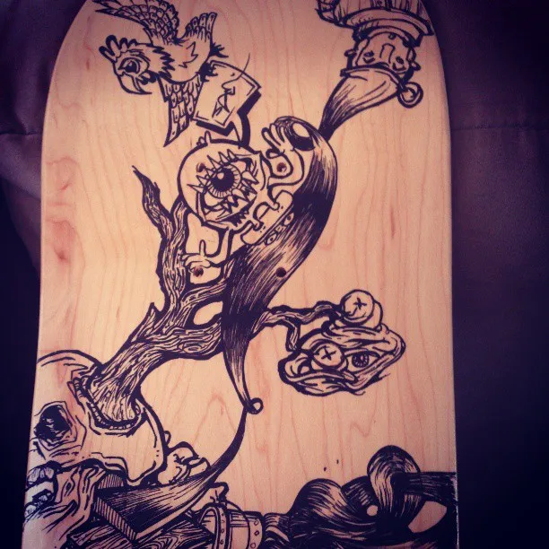 Black ink drawing on a wooden skateboard deck showing a surreal tree growing from a skull, a giant eyeball and a bird