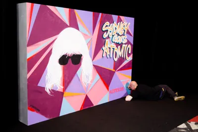 Artist painting a geometric mural of a blonde woman in sunglasses and the text Sydney Goes Atomic