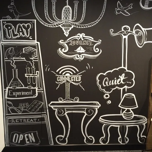 White line art mural on a black wall featuring stylized furniture, shelves, and words like Play, Journey, and Quiet