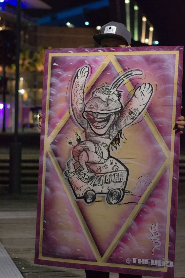 Painting of a tattooed cartoon character riding a beer van on a pink background, held by a person wearing a cap