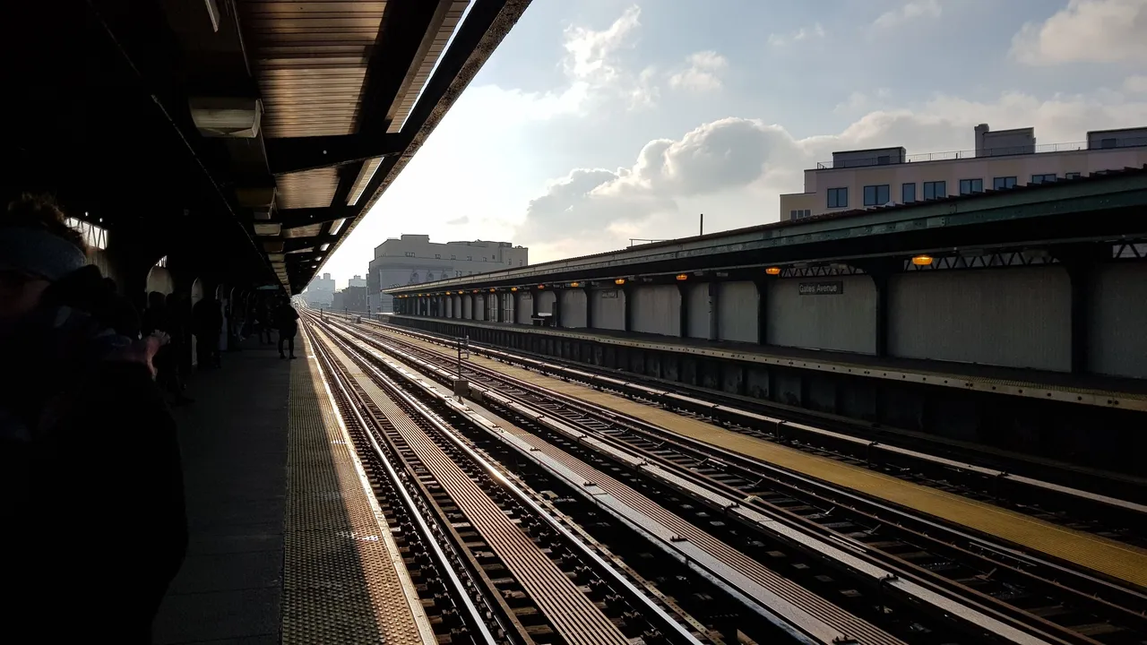 Sunlit outdoor subway platform with multiple tracks, people waiting, and a 'Gates Avenue' sign