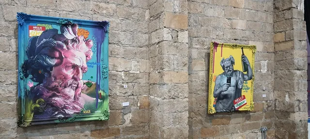 Two framed paintings on a stone wall showing classical statues remixed with neon graffiti and a smartphone selfie