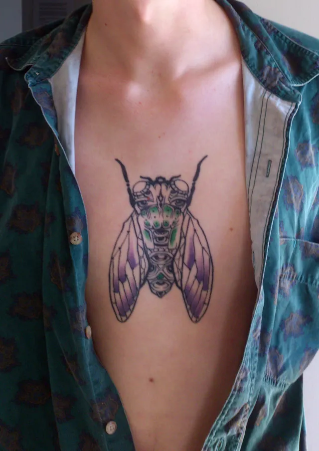 Chest tattoo of a totemic cicada featuring bold black line work, green body accents and purple wings