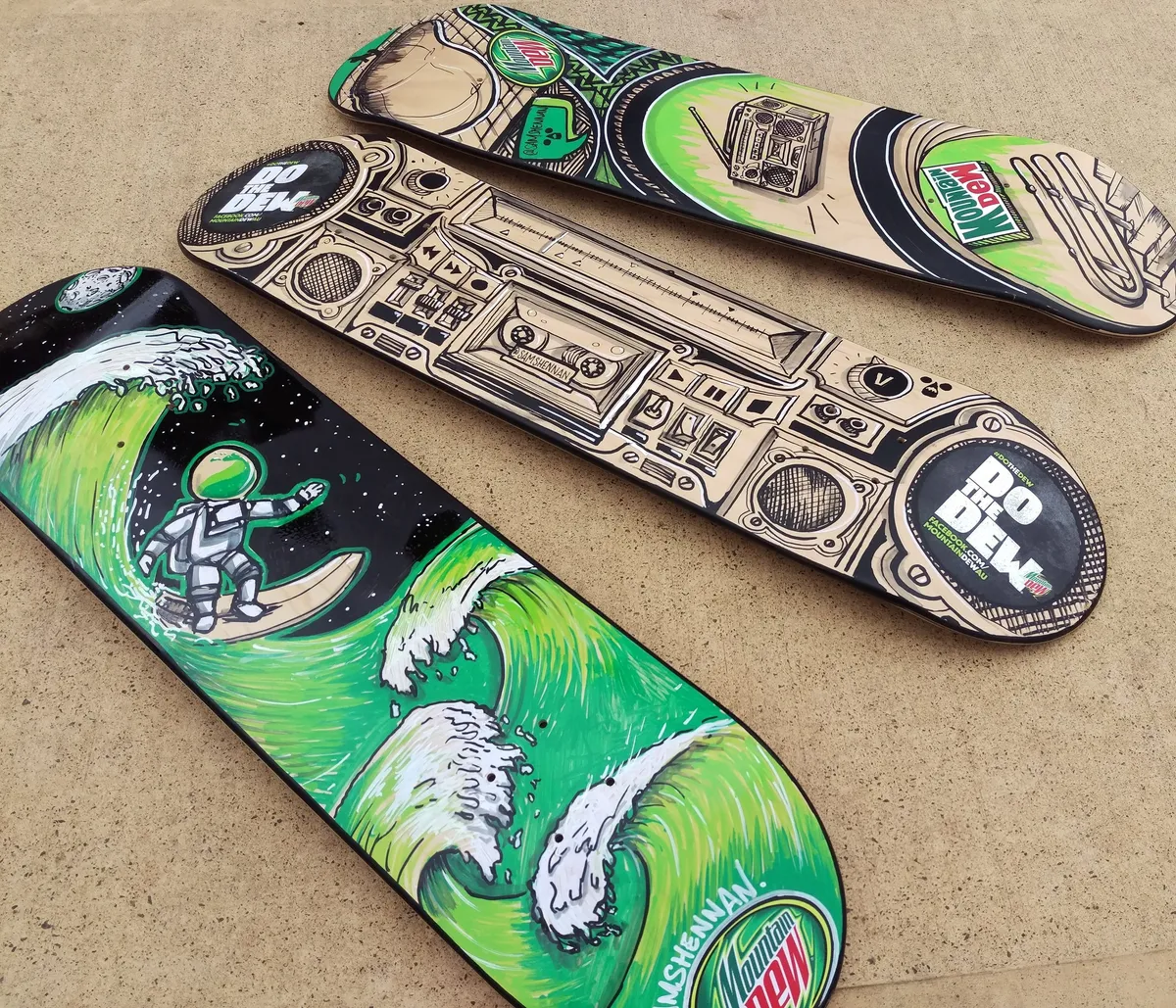 Three painted skateboards featuring a surfing astronaut on green waves, a detailed boombox, and Mountain Dew logos