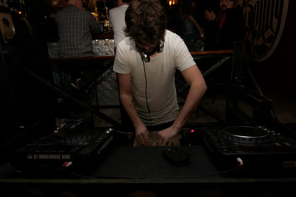 DJ in a white t-shirt operating mixing decks in a dimly lit venue