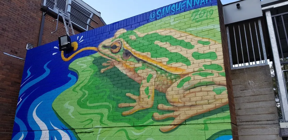 Mural of a green frog on a brick wall, its painted tongue extending to wrap around a physical floodlight fixture