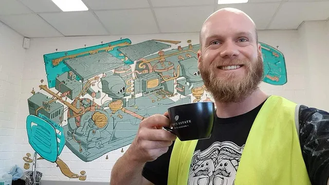 Smiling man in a high-vis vest holding a coffee cup in front of a mural of an exploded espresso machine