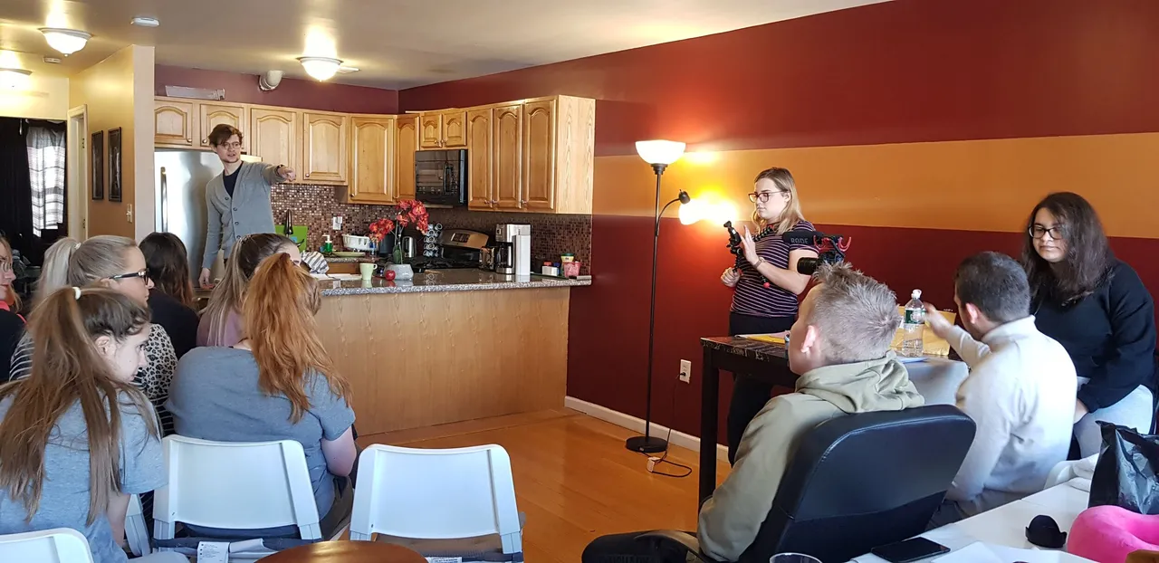 A man speaks to a group of people in a home kitchen and living room, with a woman recording