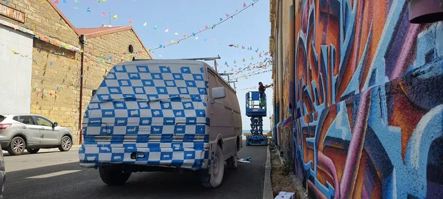 Van covered in a blue and white checkered pattern next to an artist on a lift painting a vibrant street mural