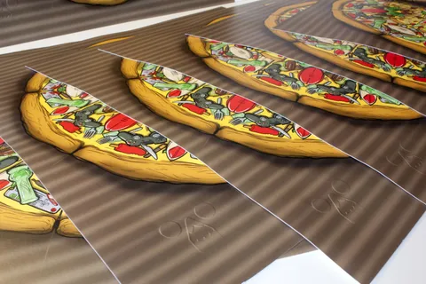 Stacked art prints of a pizza topped with game controllers and popsicles, each featuring a blind embossed logo