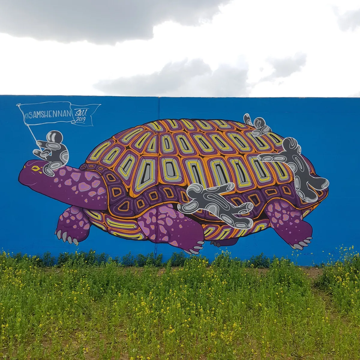 Mural on a blue wall of a giant purple tortoise with an orange patterned shell ridden by small stylized figures