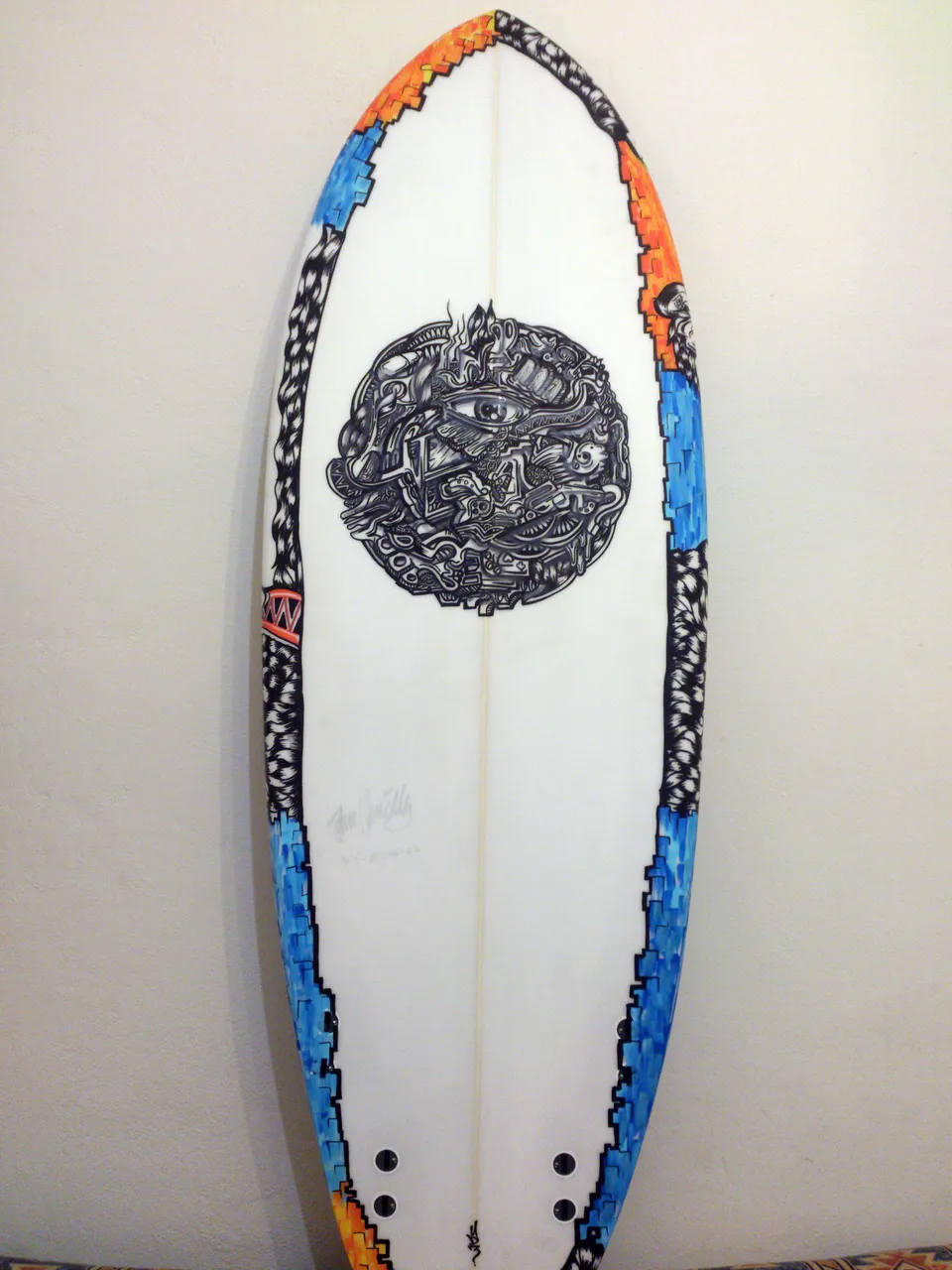 Hand-painted surfboard with a central black and white psychedelic eye motif and blue and orange patterned edges