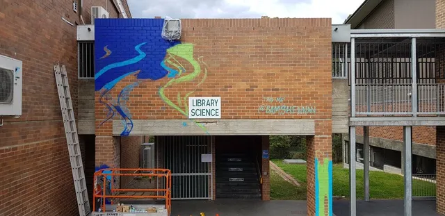 In-progress mural of abstract blue and green waves on a brick wall above a Library Science sign