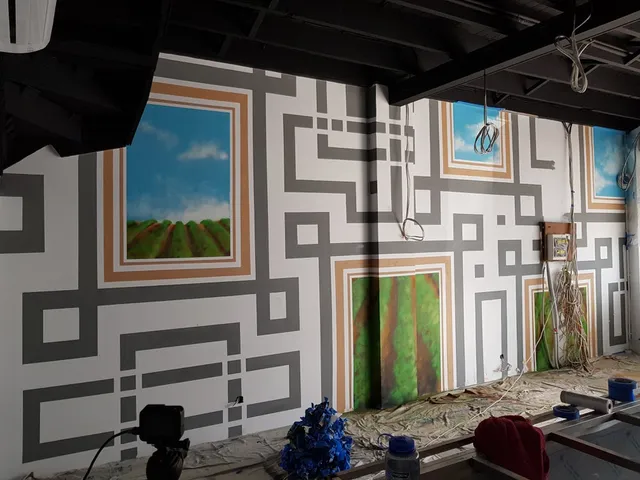 In-progress indoor mural with grey geometric lines connecting framed paintings of green fields and blue skies