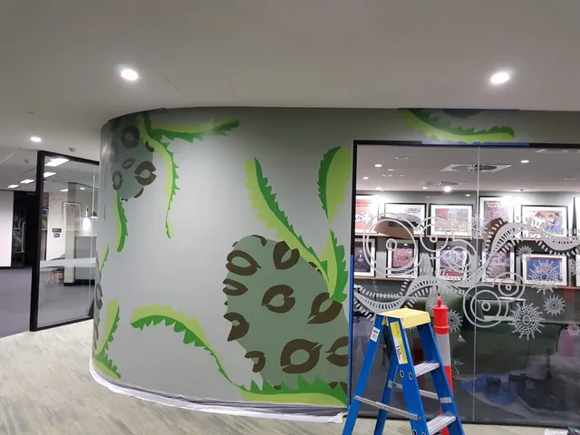 In-progress mural on a curved office wall featuring large green serrated leaves and patterned brown pods