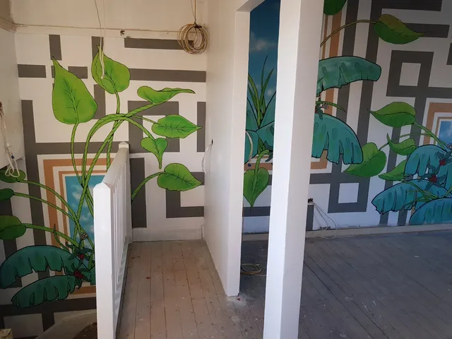 Interior mural featuring large green tropical plants and blue sky panels painted over a grey geometric grid