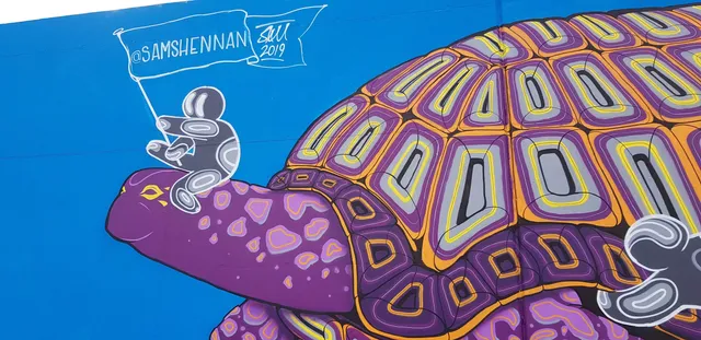 Mural of a stylized purple and orange tortoise ridden by a small figure holding a flag against a bright blue background