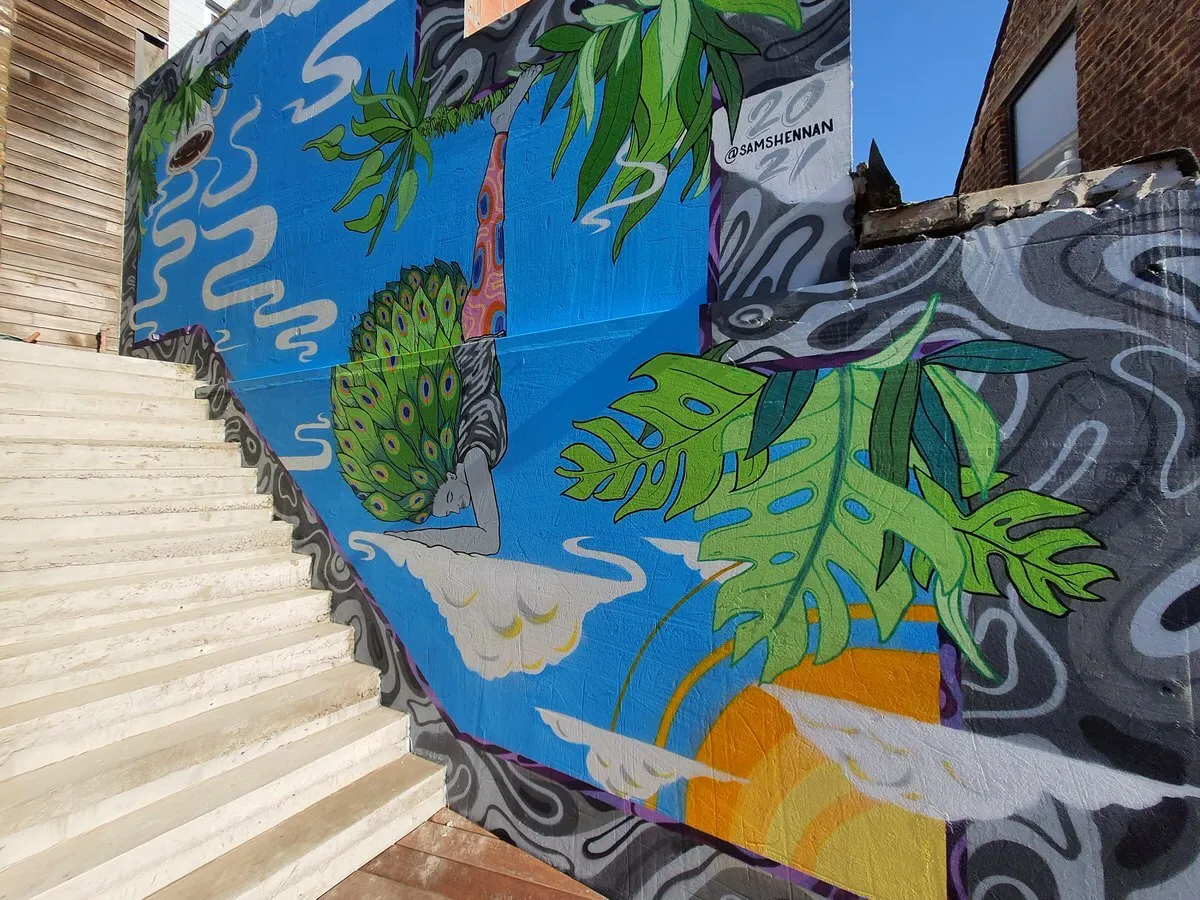 Mural of a figure handstanding on a cloud with peacock feathers, surrounded by tropical leaves on a bright blue background