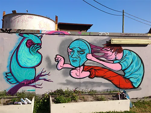 Mural of a teal owl covering its eyes next to a figure pulling a blue mask off their stretching pink face