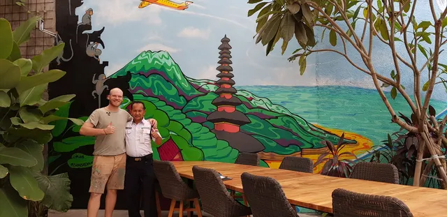 Two men giving thumbs up in front of a vibrant Bali landscape mural featuring a tiered temple, green mountains and ocean