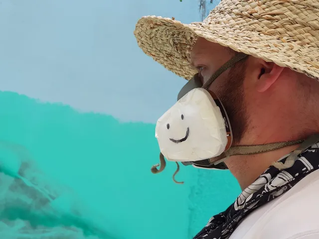 Artist in a straw hat and taped respirator mask with a drawn smiley face looking at a partially painted teal mural