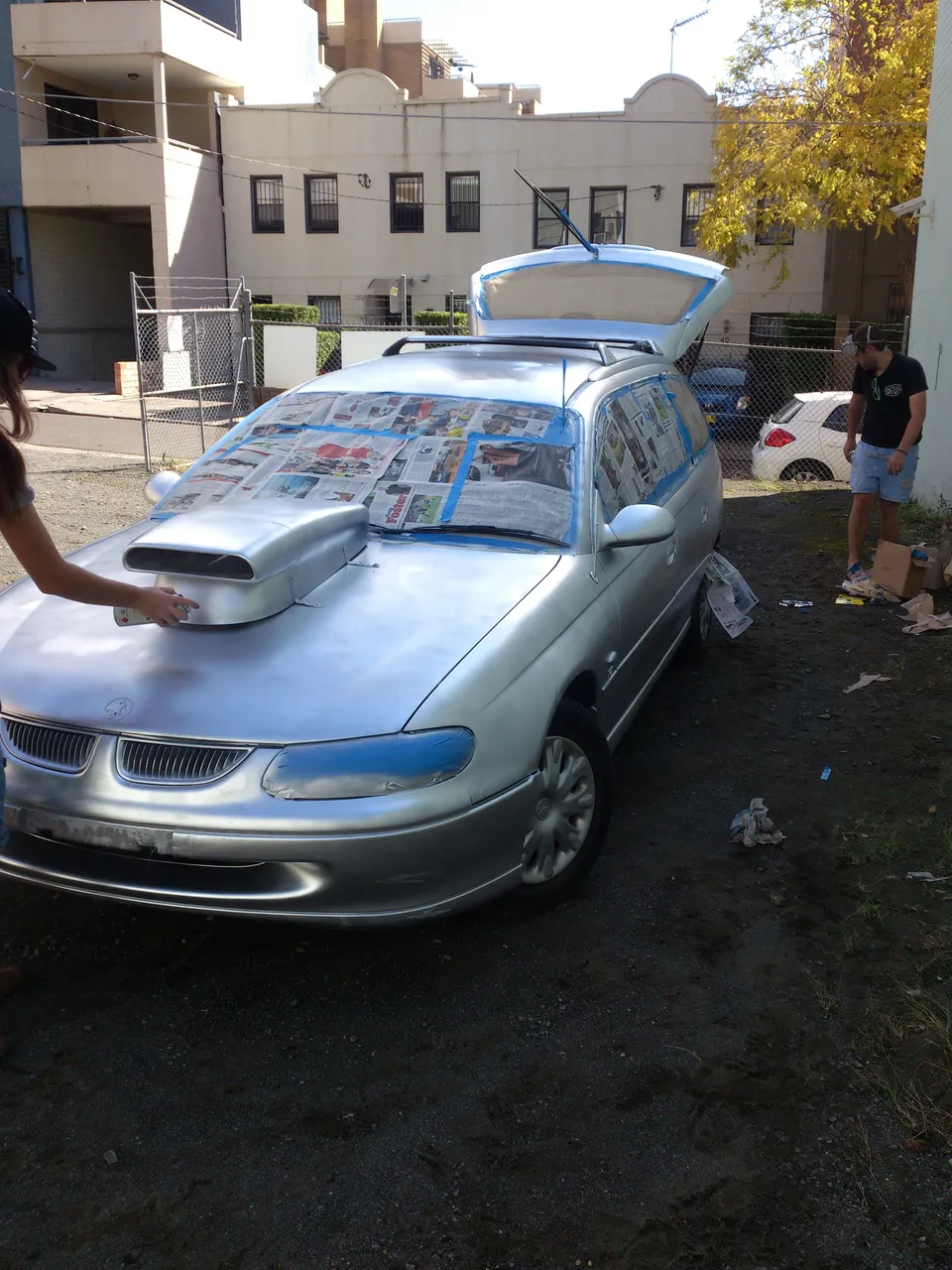 Silver station wagon with a large hood scoop and windows masked by newspaper being spray painted