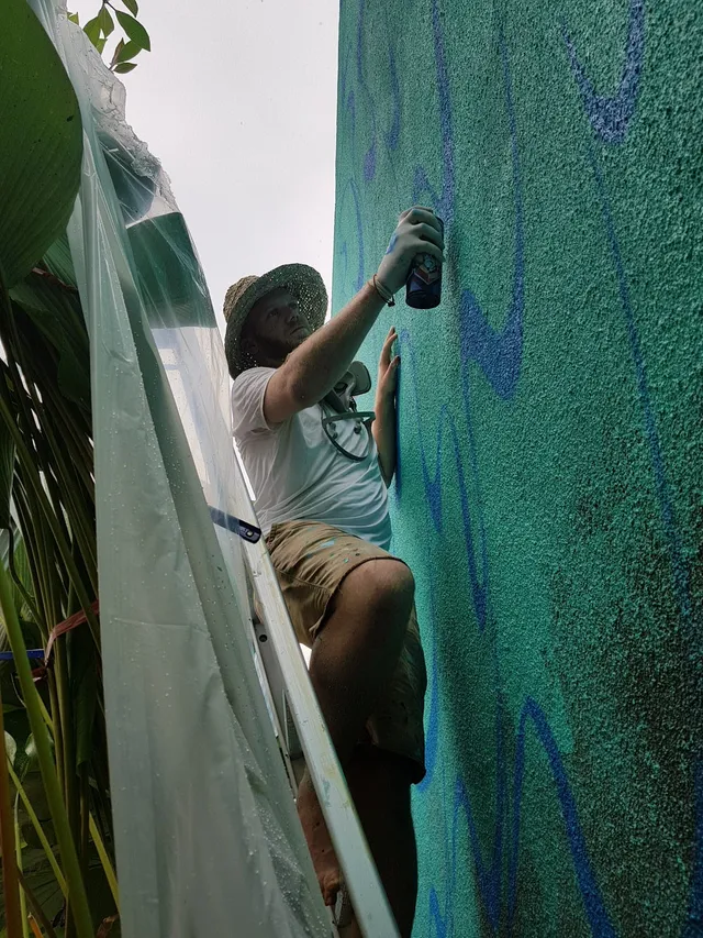 Artist on a ladder spray painting blue lines for a mural onto a textured green wall