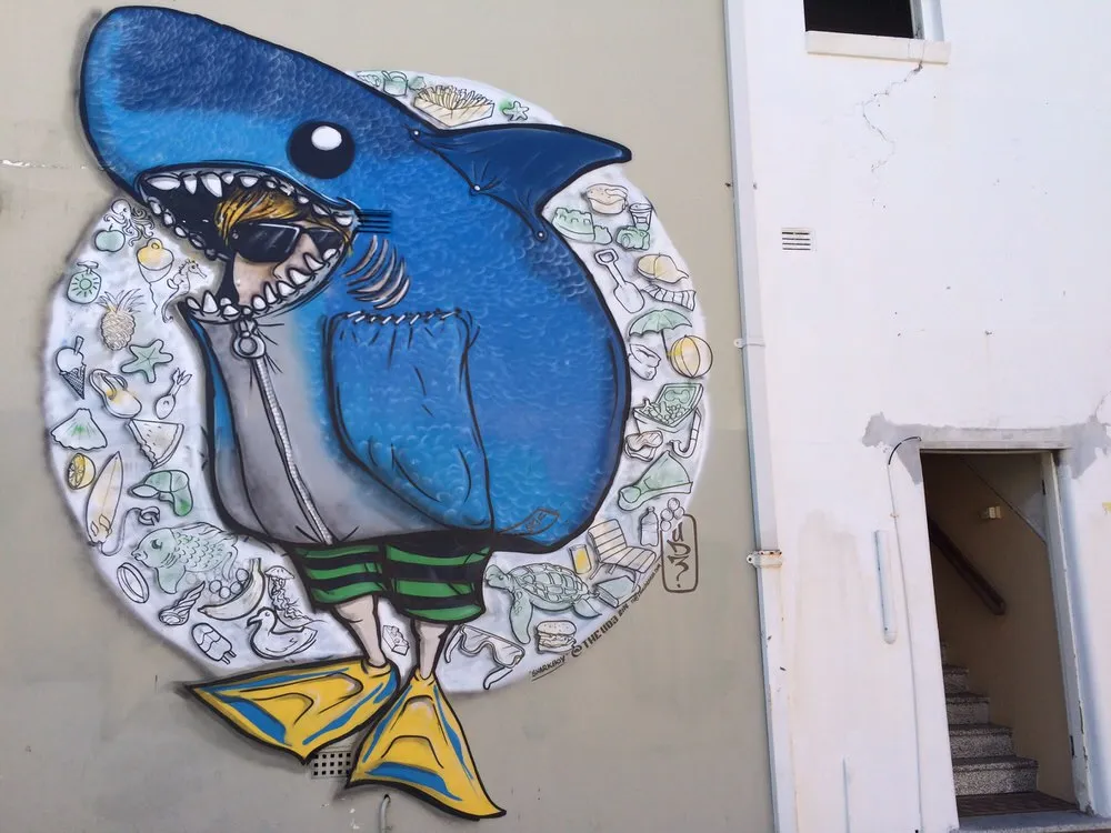 Mural of a character in a blue shark costume and yellow flippers surrounded by a circle of beach-themed line drawings
