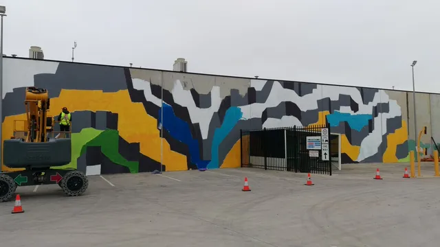 Mural in progress painted from a lift, featuring a large abstract geometric design in yellow, grey, green and blue