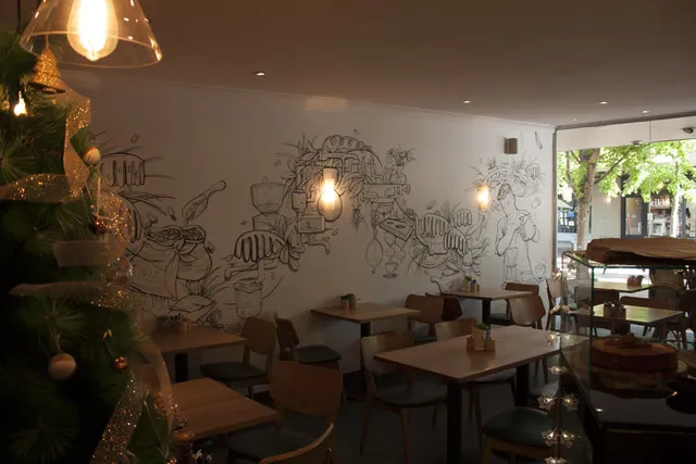 Black line mural on a cafe wall depicting intricate coffee machinery, sacks of beans, and a barista pouring a cup