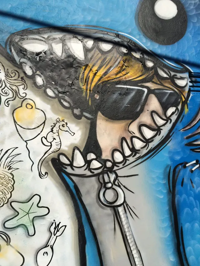 Mural detail of a blonde figure in sunglasses looking out from the zipped mouth of a blue shark suit