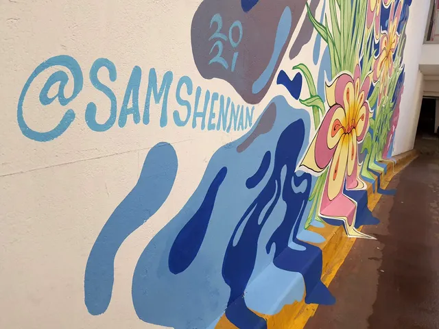 Detail of a mural on a white wall featuring abstract blue fluid shapes, pink flowers, and a painted @SAMSHENNAN handle