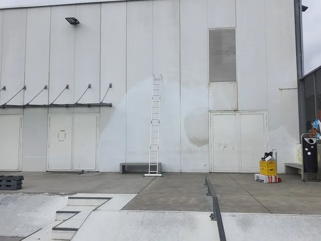 A tall ladder leaning against a blank wall at a skate park, showing a faint arched primer outline for a mural