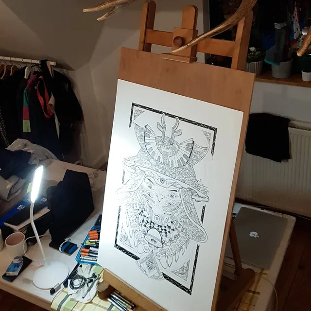 Intricate black ink drawing of a symmetrical three-eyed mask on paper resting on a wooden easel