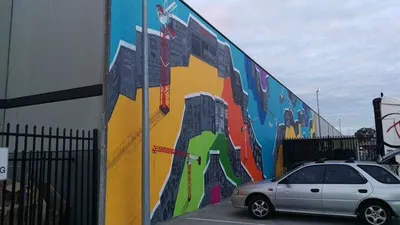 Large outdoor mural with bold geometric color blocks, dark textured structures, and red construction cranes
