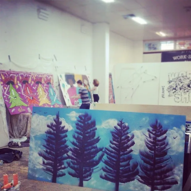 Art studio with large paintings in progress, including a foreground mural panel of four dark purple trees on a blue sky