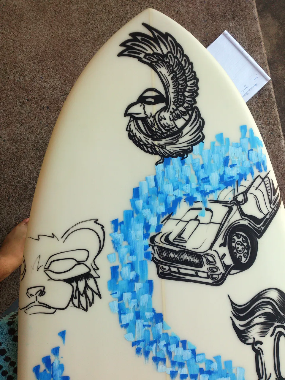 Painted surfboard with bold black line art of a bird, car, and animal face, crossed by a path of blue brushstrokes