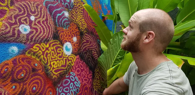 Bearded man looking at a brightly colored textured mural of intricate psychedelic patterns next to large tropical leaves