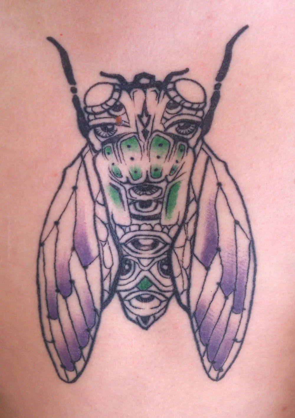Chest tattoo of a symmetrical, totemic cicada featuring multiple eyes, bold linework, green accents and purple wings
