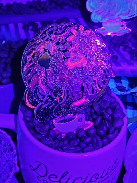 Circular enamel pin glowing under blacklight in coffee beans, showing a woman's hair rising as steam from a coffee cup