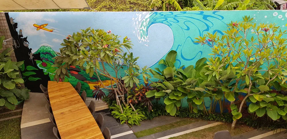Mural of a stylized blue wave, green mountains and yellow airplane on a courtyard wall behind plants and a long wooden table