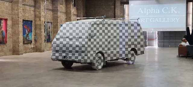 Van covered entirely in a grey and white 3D UV checkerboard pattern inside an art exhibition space