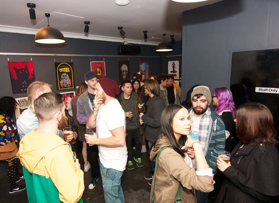 Crowd of people mingling and holding drinks at a gallery exhibition with posters displayed on the walls