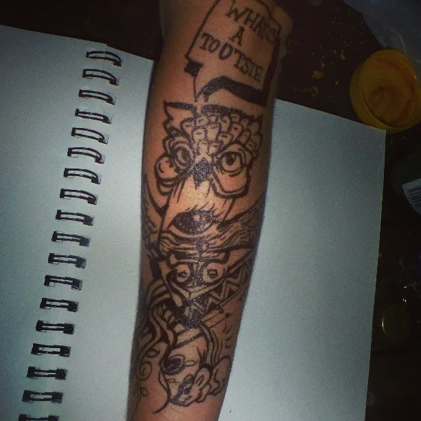 Black ink drawing on an arm featuring a totemic owl and faces with a speech bubble reading WHATS A TOOTSIE