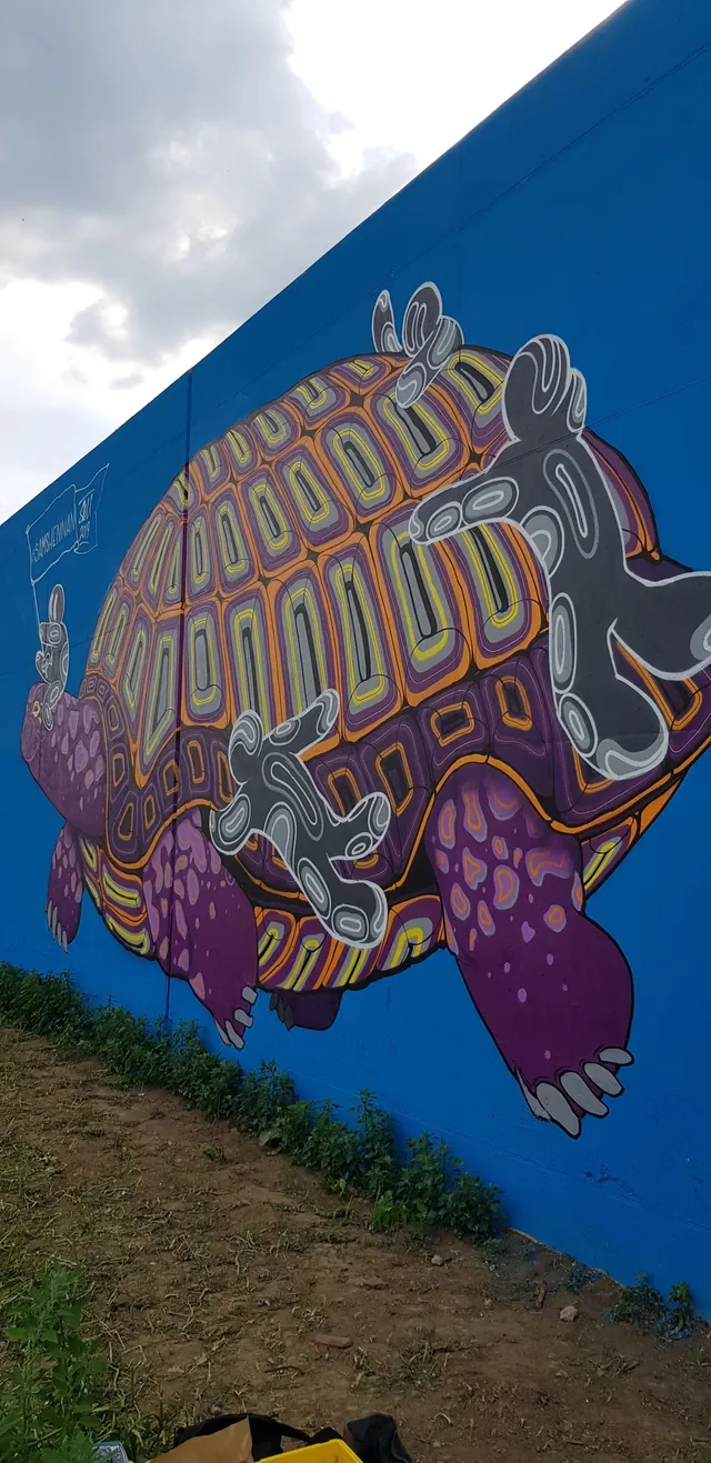 Mural on a blue wall depicting a giant patterned tortoise and floating grey totemic figures