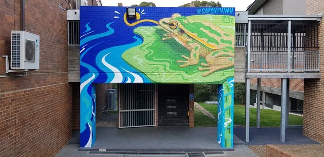 Mural on a brick wall featuring a large green frog on a lily pad beside stylized flowing blue water
