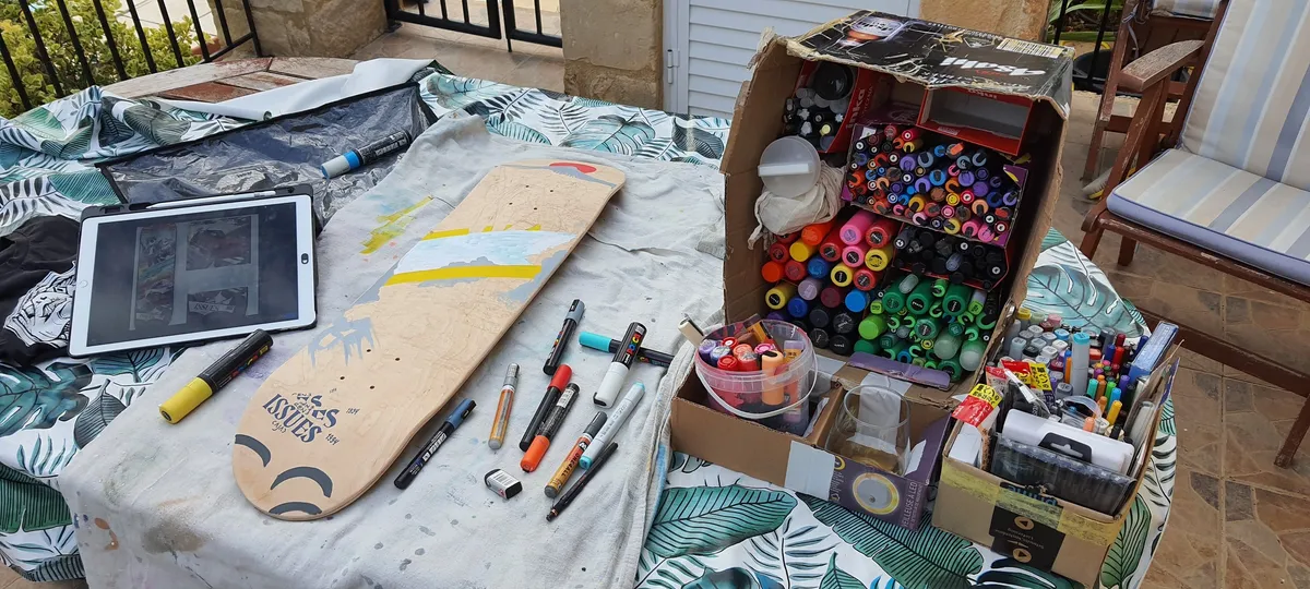 Wooden skateboard deck on a table beside an iPad and cardboard boxes packed with colorful paint markers