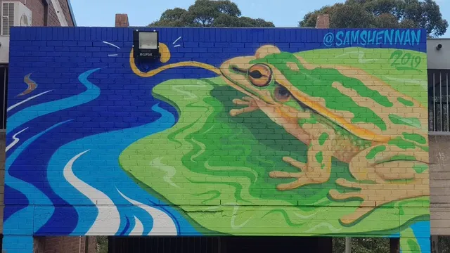 Brick wall mural of a green frog by a swirling blue river, its yellow tongue curling around a physical wall light